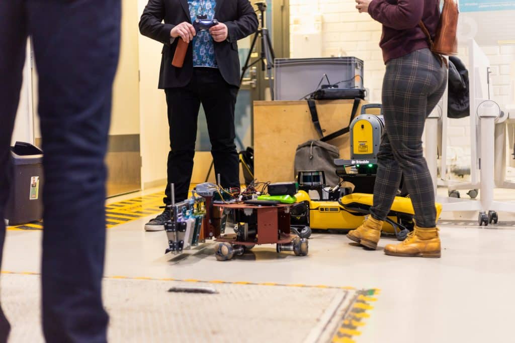 Robottidemossa pieni robotti kulkee käytävällä ja sen ympärillä on ihmisten jalkoja.